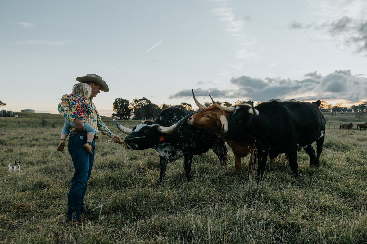 A man in an Akubra hat holding his child while patting a horned cow. 